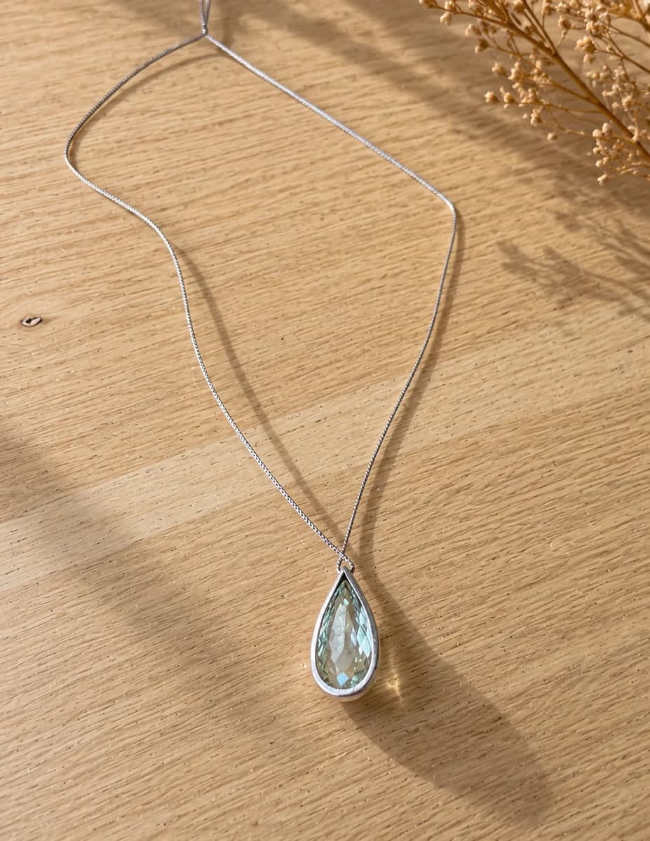 Colar de prata 950 com pingente gota de prasiolita verde-água, sobre uma mesa de madeira clara com flores secas ao fundo.